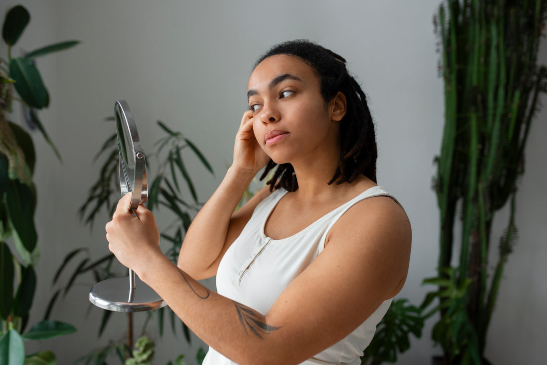 Mujer mexicana con piel morena con un fondo blanco y plantas verdes y mirandose en un espejo realizandoce una rutina de skincare con los productos de Tezzalli